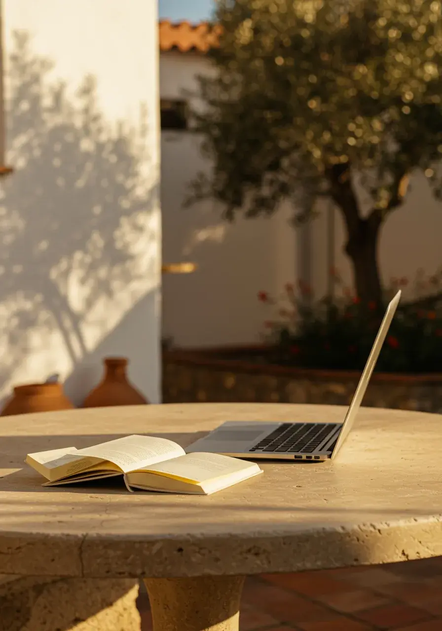 Otwarta książka i laptop na wapiennym stole na dziedzińcu w ciepłym śródziemnomorskim świetle popołudniowym, drzewo oliwne w tle